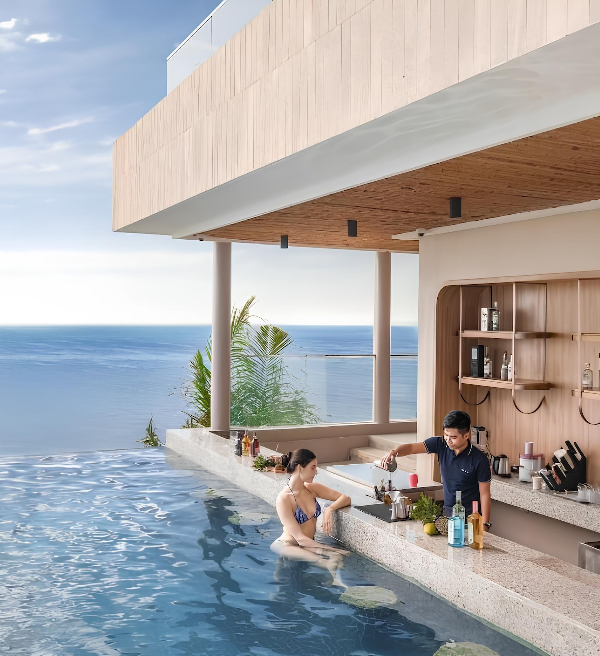 An infinity pool extends towards a serene ocean view, complemented by a wet bar featuring an array of beverages and glassware. One guest is seated in the water, while another prepares a drink. Soft lighting contributes to the relaxing ambiance.
