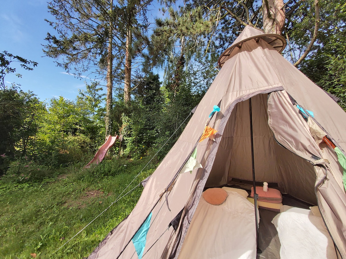 A spacious tent is surrounded by greenery, with colorful decorative elements adorning the entrance. In the background, a small slide is visible amidst the trees, providing a playful outdoor feature. The interior features two beds with minimal furnishings, creating a simple and inviting atmosphere.