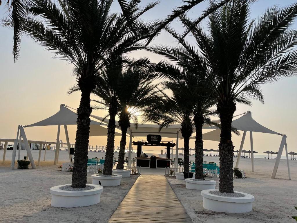 A sandy beach area is lined with tall palm trees, leading to a shaded refreshment area. Comfortable turquoise seating is visible beneath a large canopy, providing a relaxed atmosphere. The horizon is softly illuminated by a setting sun, creating a serene backdrop for relaxation.