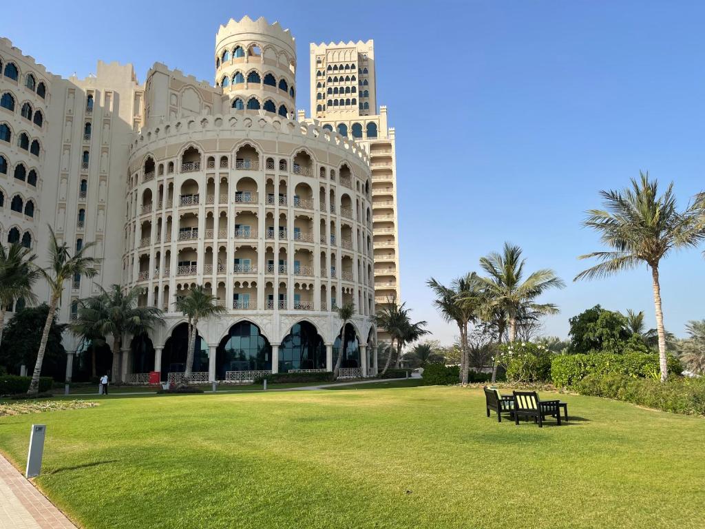 A grand building features an ornate facade with multiple arches and balconies, surrounded by a lush green lawn. Palm trees are positioned throughout the area, providing shade and enhancing the serene ambiance. A pair of black benches offers seating for relaxation amidst the natural landscape.