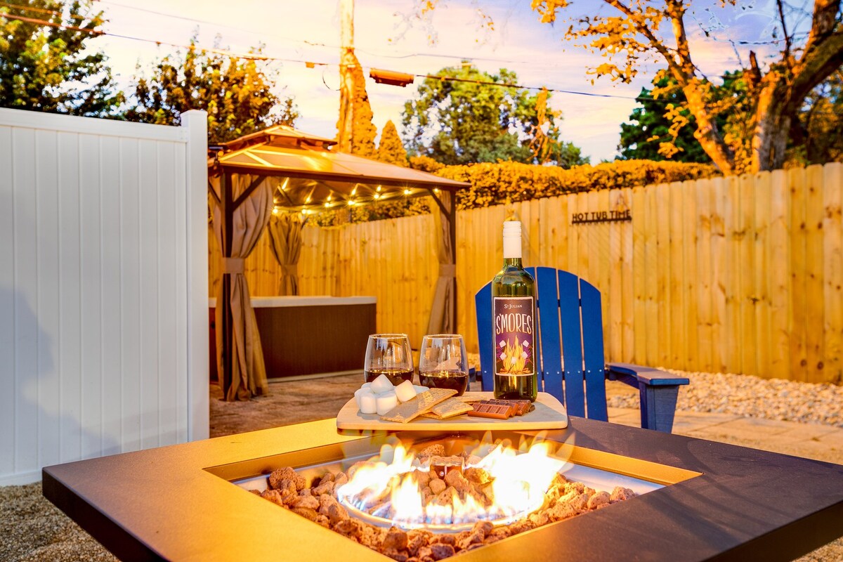A backyard space features a cozy fire pit surrounded by pebbles, with flames dancing above. A wooden tray holds wine glasses, a wine bottle, and s'mores ingredients. A pergola sits in the distance, and a hot tub is visible, framed by warm lighting and privacy fencing.