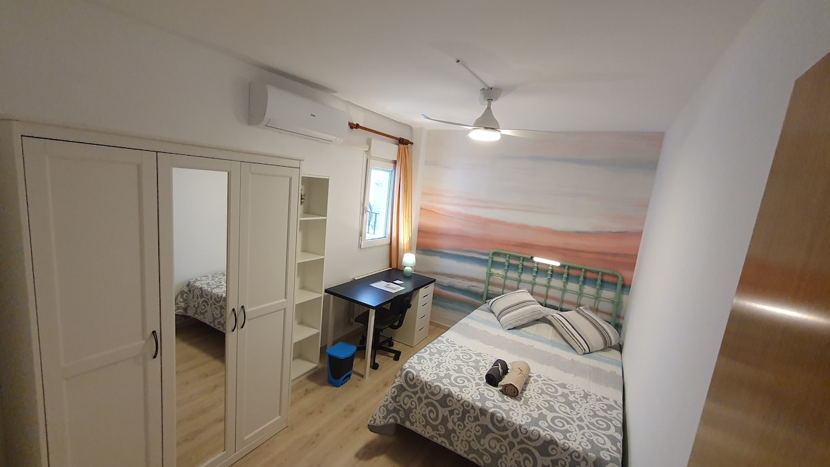 A modern bedroom features a large bed with patterned bedding and decorative pillows. A study area is equipped with a desk and lamp, positioned near a window. A spacious wardrobe with mirrored doors stands against one wall, complemented by light wooden flooring and soft lighting.