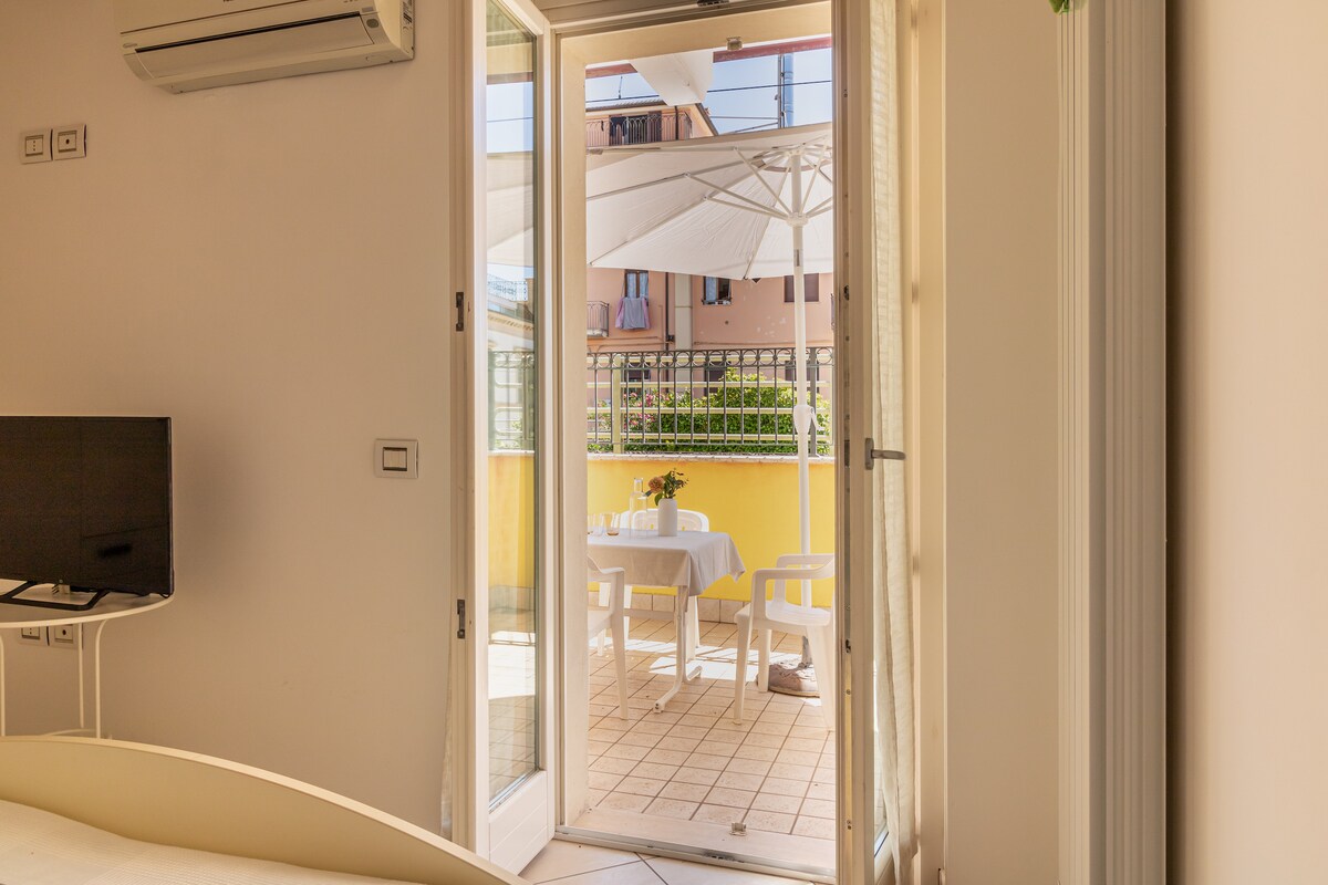 A bright indoor space features glass doors opening onto a terrace, providing access to outdoor seating and a table set for dining. Sunlight streams in, highlighting the fresh decor and climate control unit mounted on the wall.