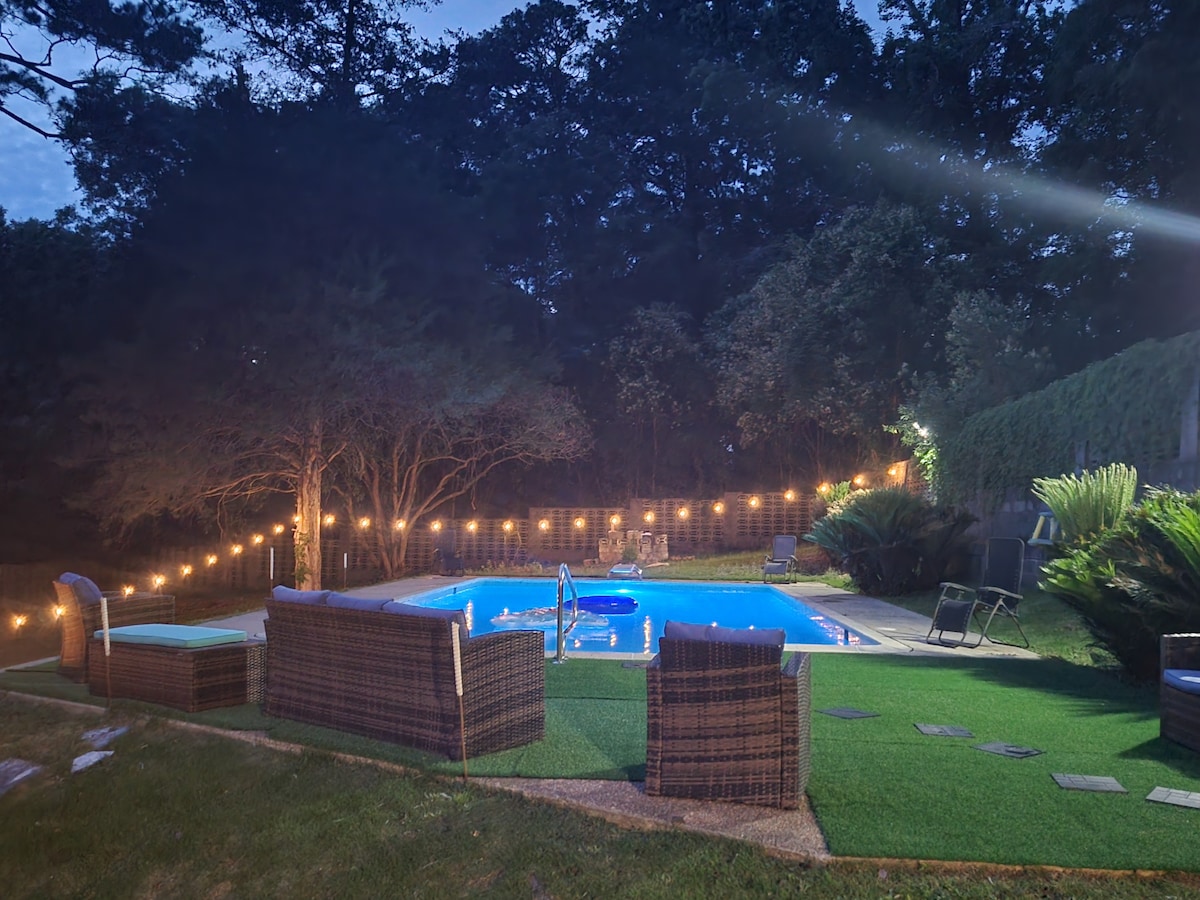 A serene outdoor space is illuminated by string lights, creating a cozy ambiance around a turquoise pool. Several cushioned lounge chairs are arranged nearby on lush green grass, surrounded by trees, offering a private retreat for relaxation.