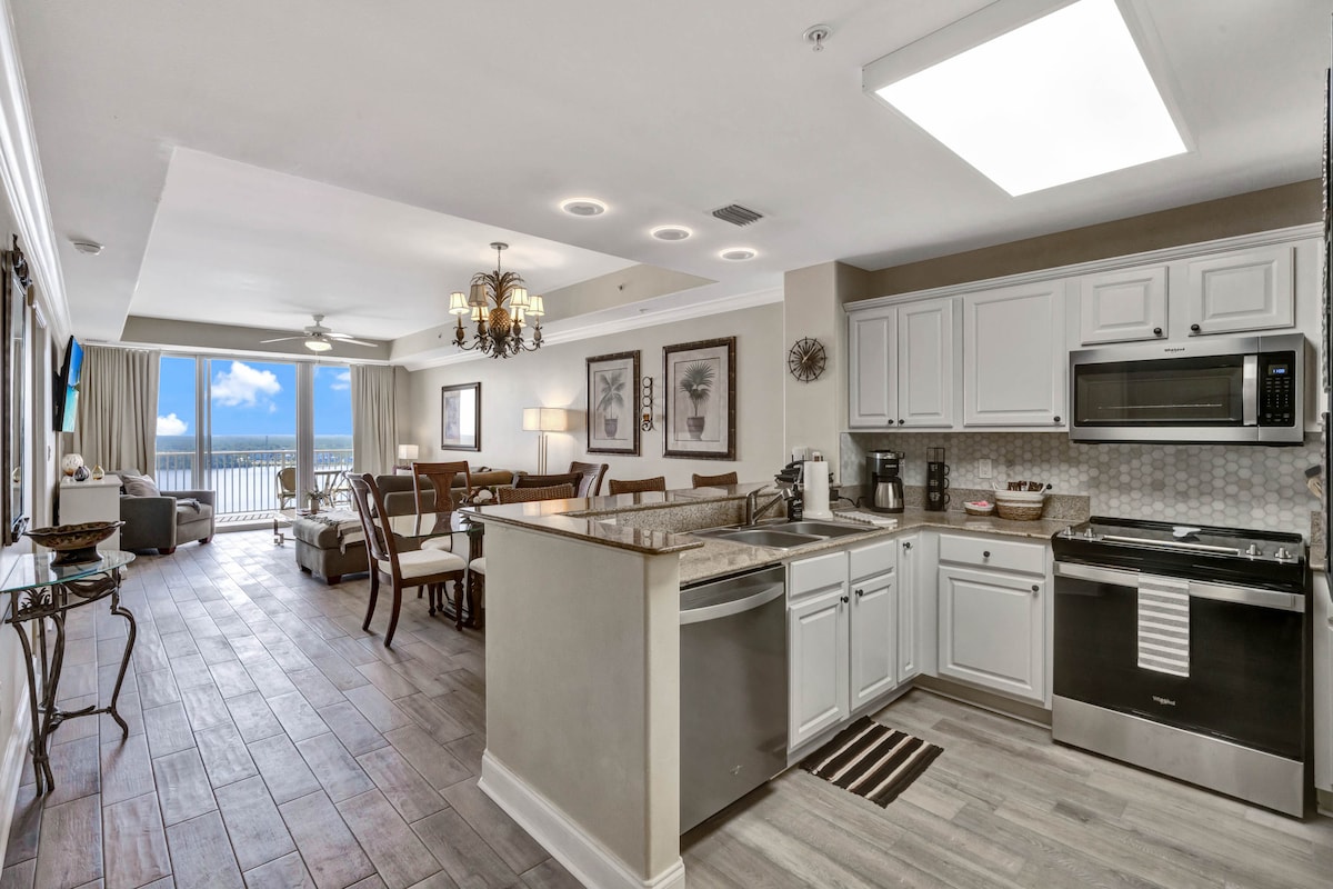 A modern kitchen is equipped with stainless steel appliances, including a microwave and dishwasher, set against white cabinetry. Natural light enters from the spacious living area, which features a comfortable seating arrangement and a view of the lakefront balcony through large glass doors.