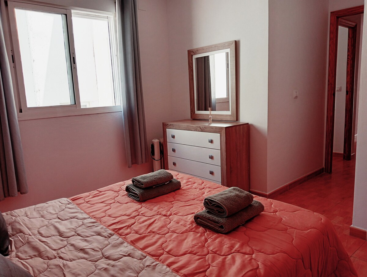 A bright sleeping area is equipped with a spacious bed layered with a coral-colored quilt. Two neatly rolled towels are placed on the bed. A dresser with four drawers is positioned nearby, complemented by a mirror, while natural light filters through a window covered by light drapes.