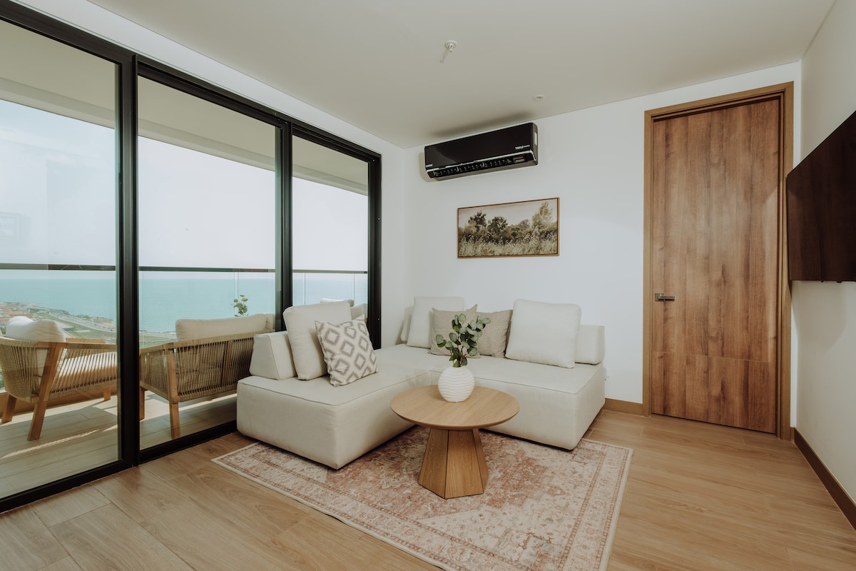 A bright living area features a modern sofa set with neutral tones and a wooden coffee table. Large glass doors provide a view of the ocean and open to a balcony. Soft natural light illuminates the space, complementing the warm wood flooring.