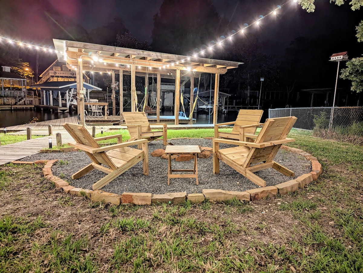A cozy outdoor seating area is highlighted, featuring four wooden Adirondack chairs arranged around a central table. Soft lighting from overhead string lights creates a warm ambiance, illuminating the space near the water. The gravel surface is surrounded by a circular stone border.