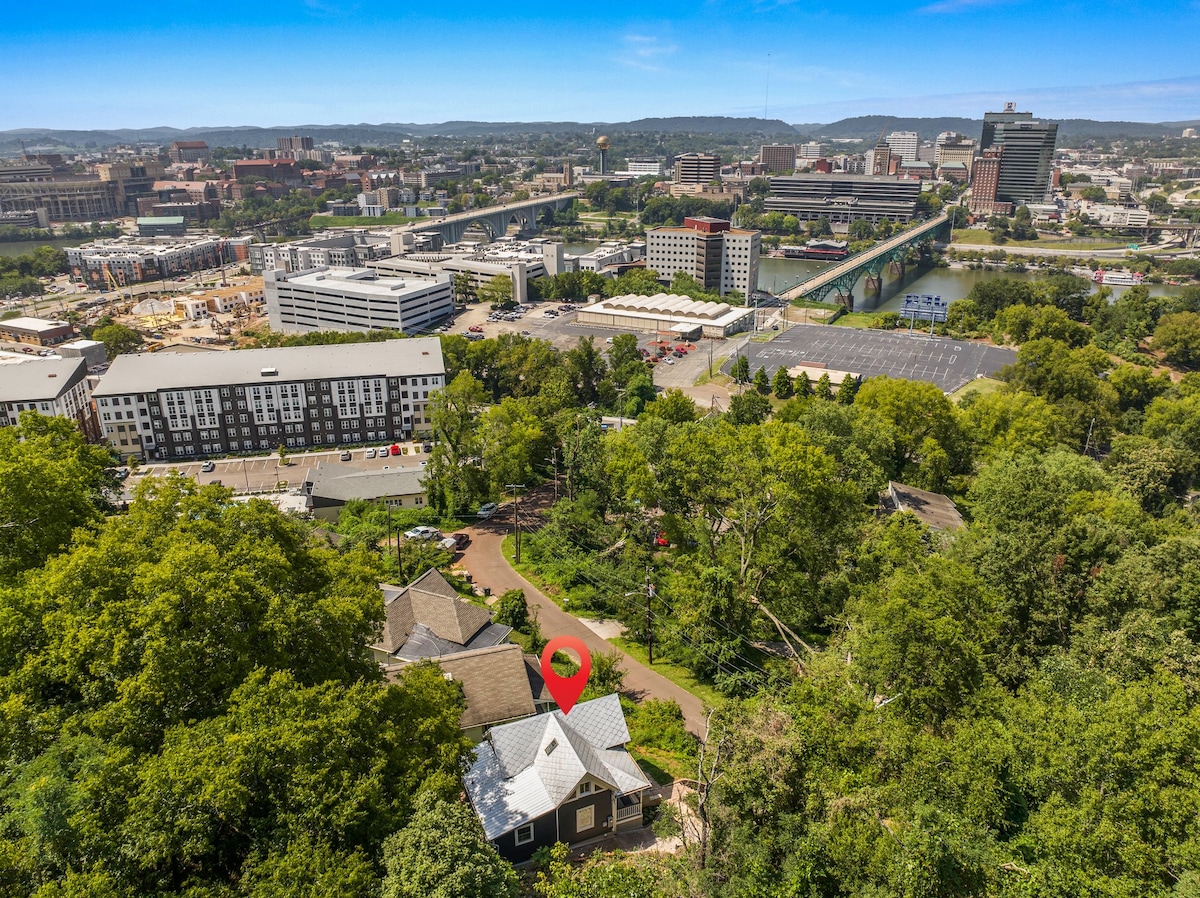 An aerial view reveals a historic home nestled among trees on a hill, overlooking downtown Knoxville and the Tennessee River. The cityscape features a mix of buildings and bridges in the distance, with greenery surrounding the property, emphasizing its serene location.