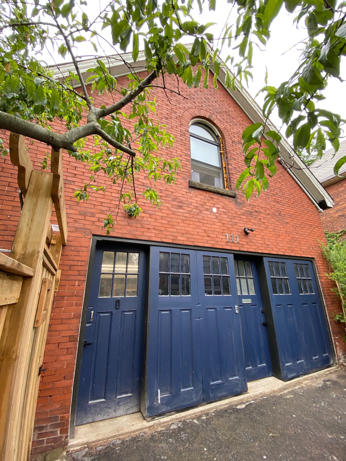 The exterior of a brick building features double doors painted in a deep blue hue, complemented by multiple windows above. The upper arch of the building is accentuated by a rounded window, while lush greenery partially frames the entrance, enhancing the inviting appearance.