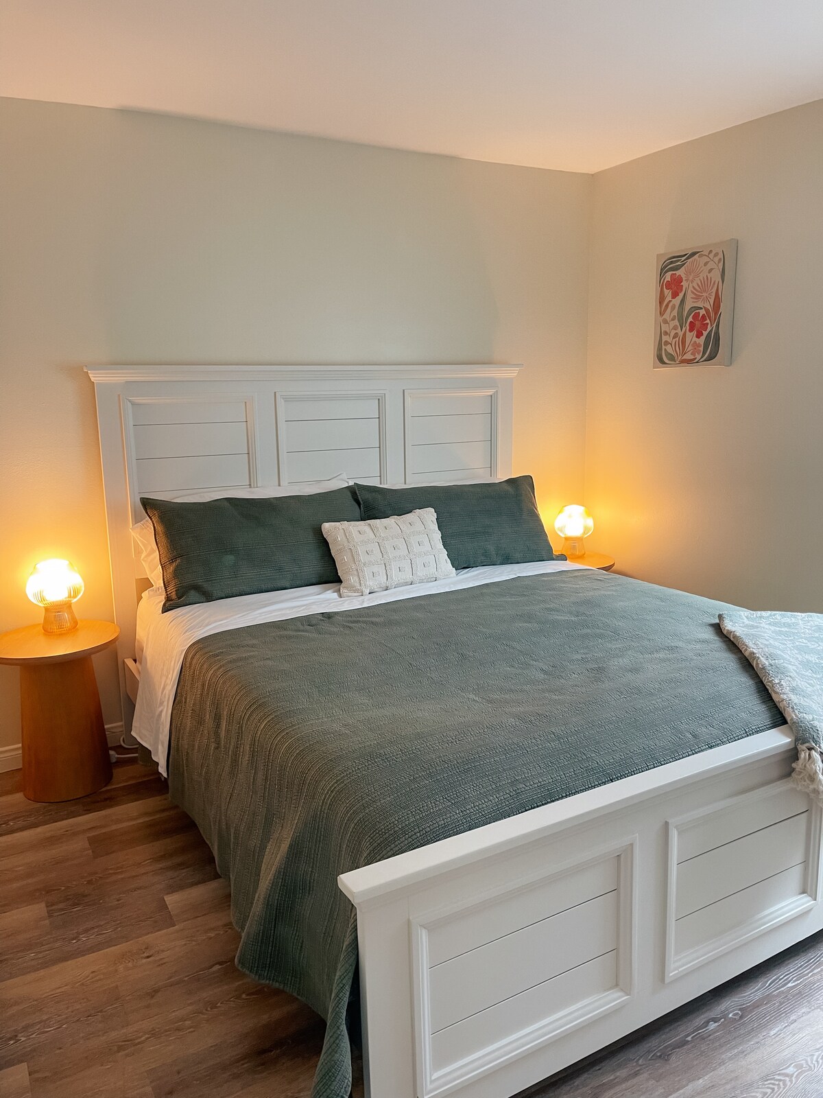 A calming bedroom is furnished with a spacious bed dressed in soft green bedding. Nightstands with warm lighting flank each side of the bed. A piece of wall art adds a touch of color, while natural light enhances the serene atmosphere.