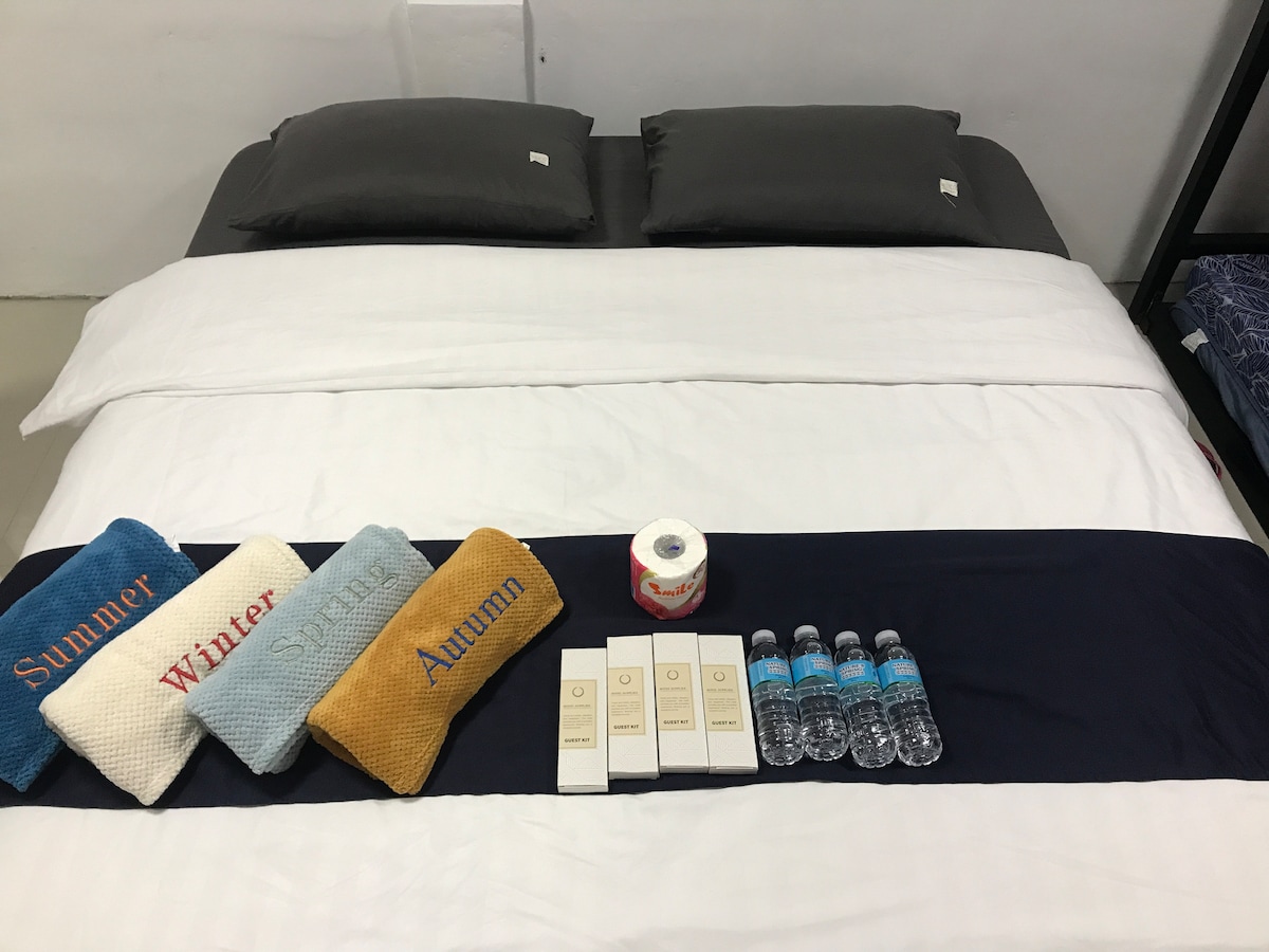 A spacious bed is neatly arranged with a crisp white duvet and dark gray pillows. Decoratively placed along the edge are three colorful cushions labeled 'Summer,' 'Winter,' and 'Autumn.' Bottled water and toiletries are aligned on the bedspread, offering convenience for guests.