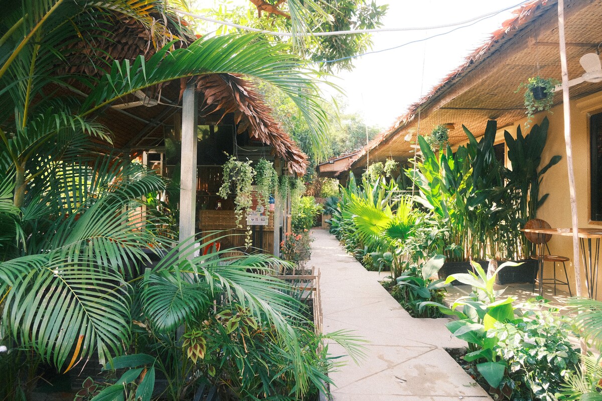 A stone pathway winds through a lush tropical garden, flanked by a variety of vibrant green plants and trees. Rustic structures with thatched roofs are visible in the background, providing a serene and natural setting for guests to explore.