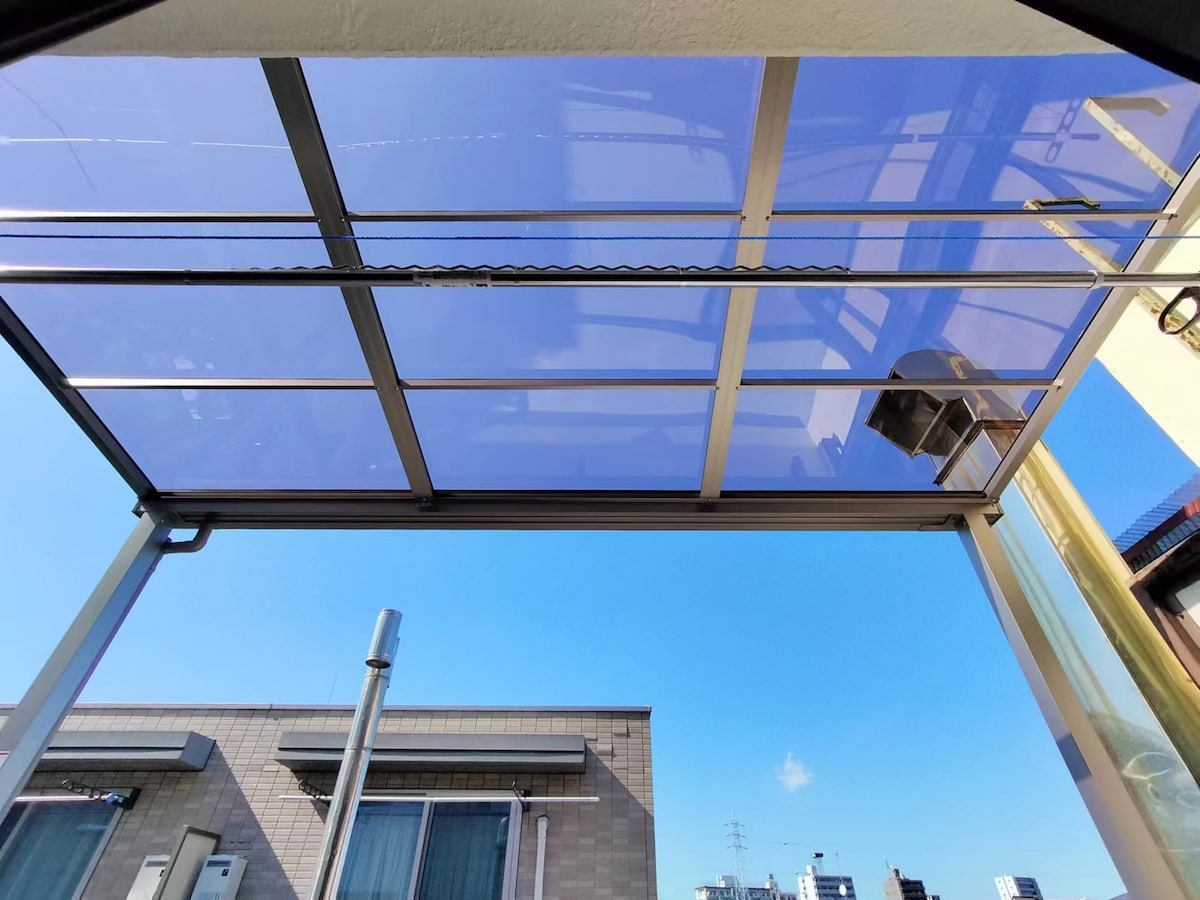 A clear covered terrace is seen from below, showcasing a transparent roof that allows sunlight to filter through. Blue skies are visible above, contributing to a bright, open atmosphere. Surrounding buildings are partially visible, providing context to the outdoor space.