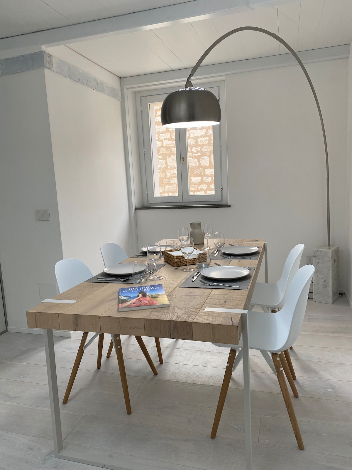 A dining area features a wooden table with a natural finish, accompanied by four white chairs. A stylish floor lamp arcs gracefully above. The table is set with plates and glassware, while a decorative basket adds a subtle touch. Light enters through a nearby window.