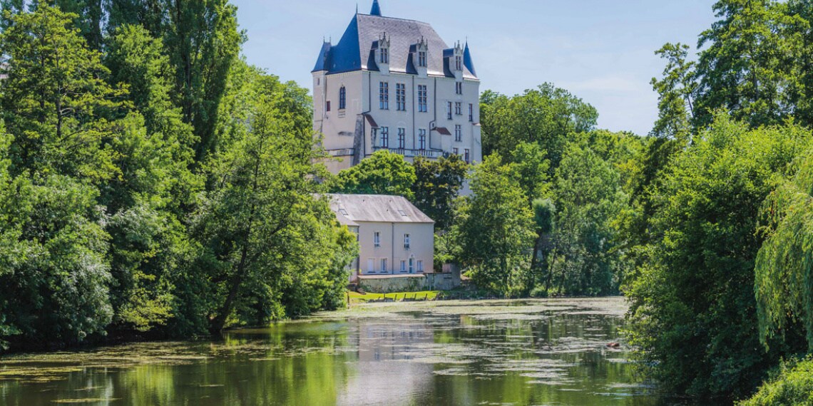 A serene scene featuring a grand building nestled among lush green trees, reflected in a calm body of water. The surrounding landscape is rich with foliage, creating a peaceful atmosphere, while sunlight bathes the area in warmth.