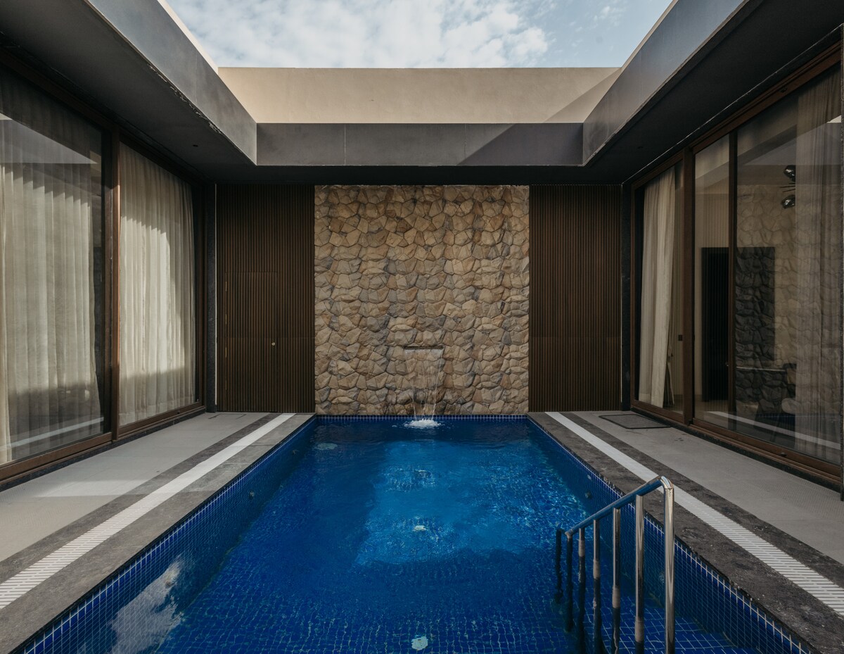 A serene pool area is framed by large glass doors, allowing natural light to fill the space. Smooth stone walls are complemented by a calming water feature at the far end of the pool. A sleek stairs leads into the refreshing blue water.