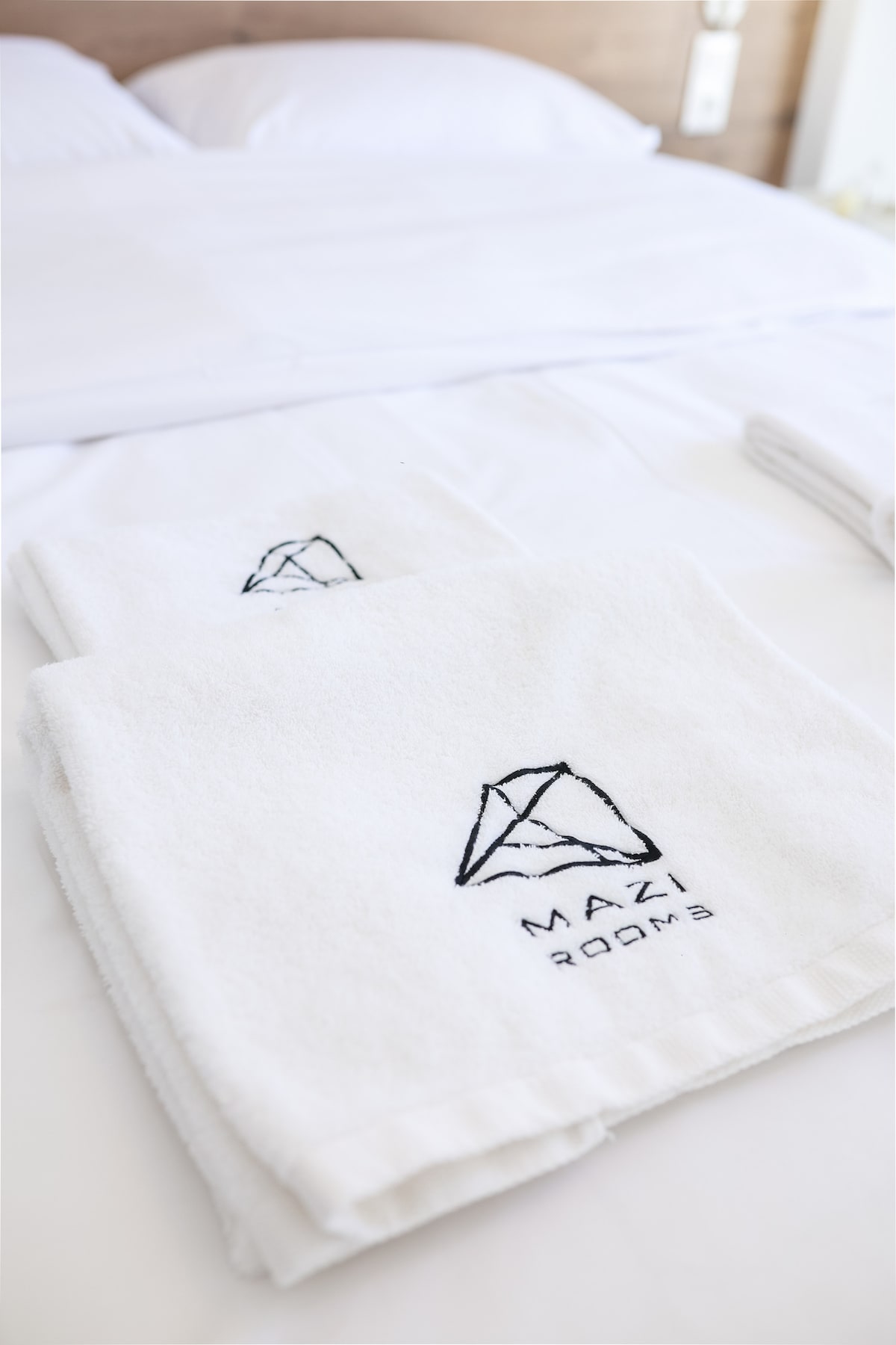 Two neatly folded towels with a logo are placed on a white bedspread, highlighting the clean and organized presentation of the sleeping area. The bed is adorned with crisp white linens, creating a fresh and inviting environment.