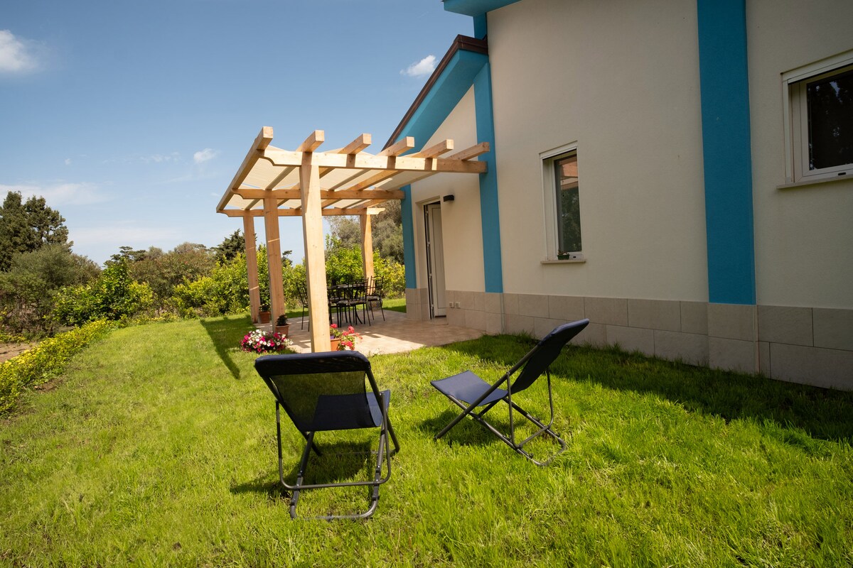A well-maintained outdoor area features a sunshade pergola with seating. Two lounge chairs are positioned on the green lawn, inviting relaxation. Surrounding greenery and flowering plants add a touch of color to the serene garden space adjacent to the property.