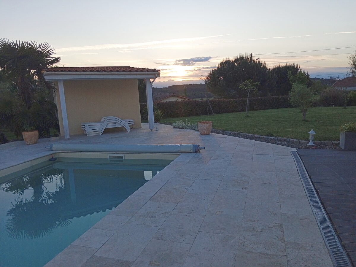 The image showcases a serene outdoor space featuring a rectangular swimming pool, its surface reflecting the soft colors of the sunset. A snug seating area with lounge chairs sits under a covered patio. Lush greenery and well-maintained gardens surround the pool area.