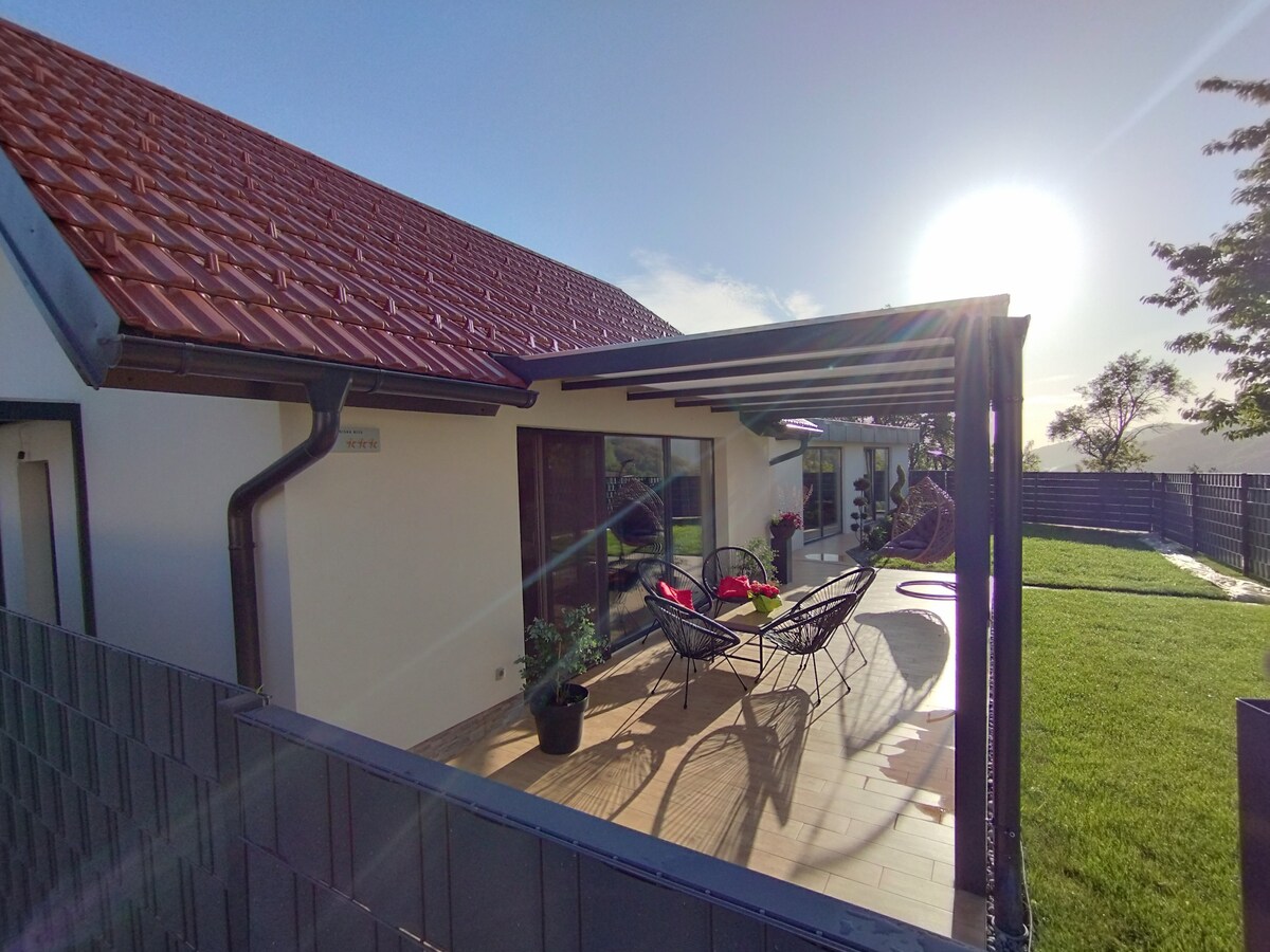 The exterior of the holiday house is shown, featuring a covered terrace with modern seating. Sunlight illuminates the area, highlighting the green lawn and surrounding landscape. The building exhibits a sloped roof and large glass doors that provide a view of the outdoor space.