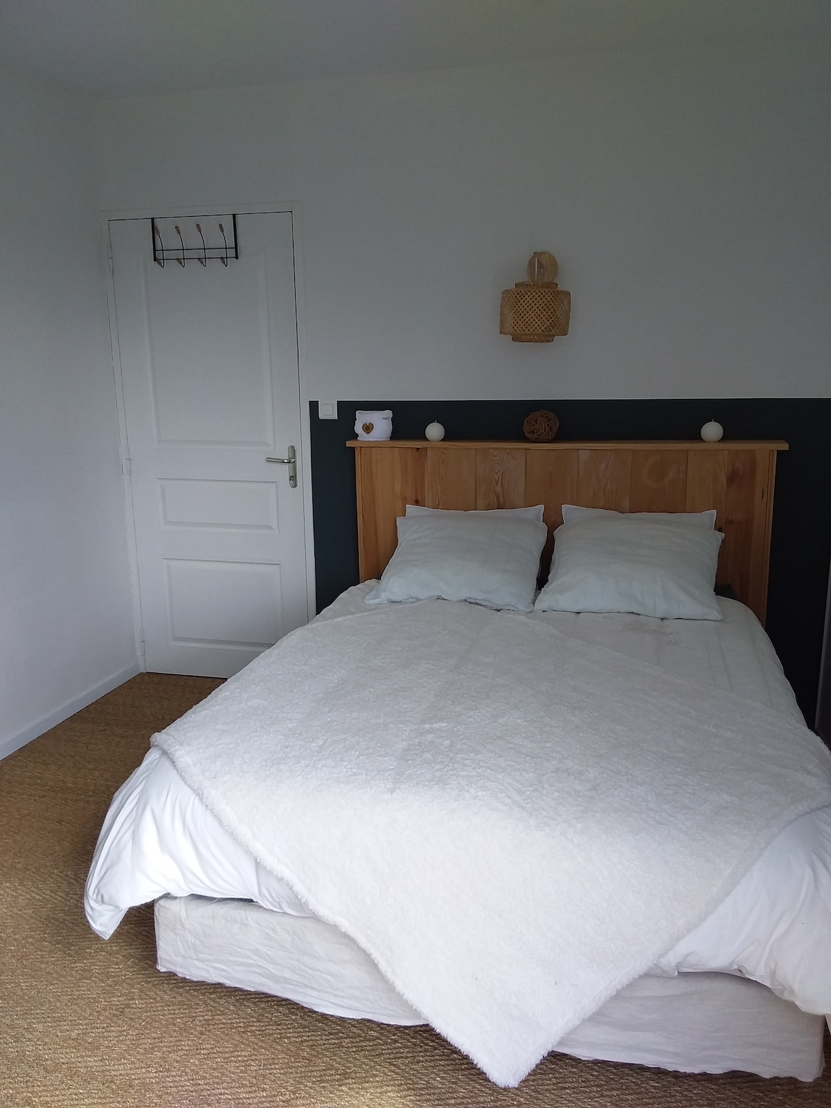 A simple bedroom is presented, featuring a spacious bed dressed in white linens and two plush pillows. The light-colored walls highlight a wooden headboard. A small decorative piece and a mirror are mounted above the bed, with a door leading to another room visible in the background.