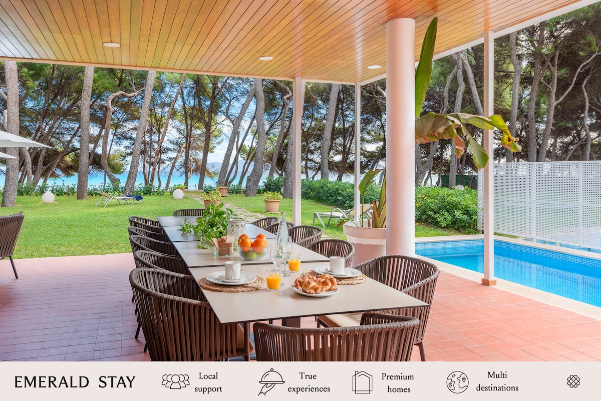 An outdoor dining area is highlighted under a wooden ceiling, featuring a long table set with fruit, pastries, and drinks. Lush greenery surrounds the space, with a view of the serene pool and beach visible through large windows.