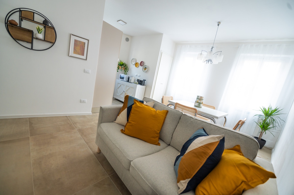 The spacious living area features a light-colored sofa adorned with cushions in shades of yellow and blue. A dining table with wooden chairs is visible in the background, alongside a well-equipped kitchenette. Natural light floods in through large windows, enhancing the bright ambiance.