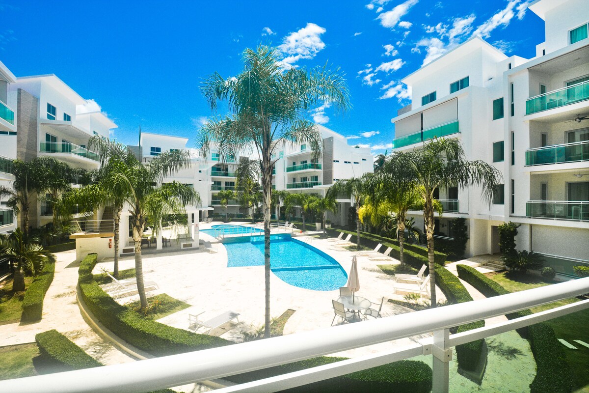 The image captures a vibrant community pool area surrounded by lush palm trees and manicured green spaces. Clear blue skies are visible overhead, while the pool features comfortable lounge chairs and an inviting swimming area, highlighting the tranquil ambiance of the complex.