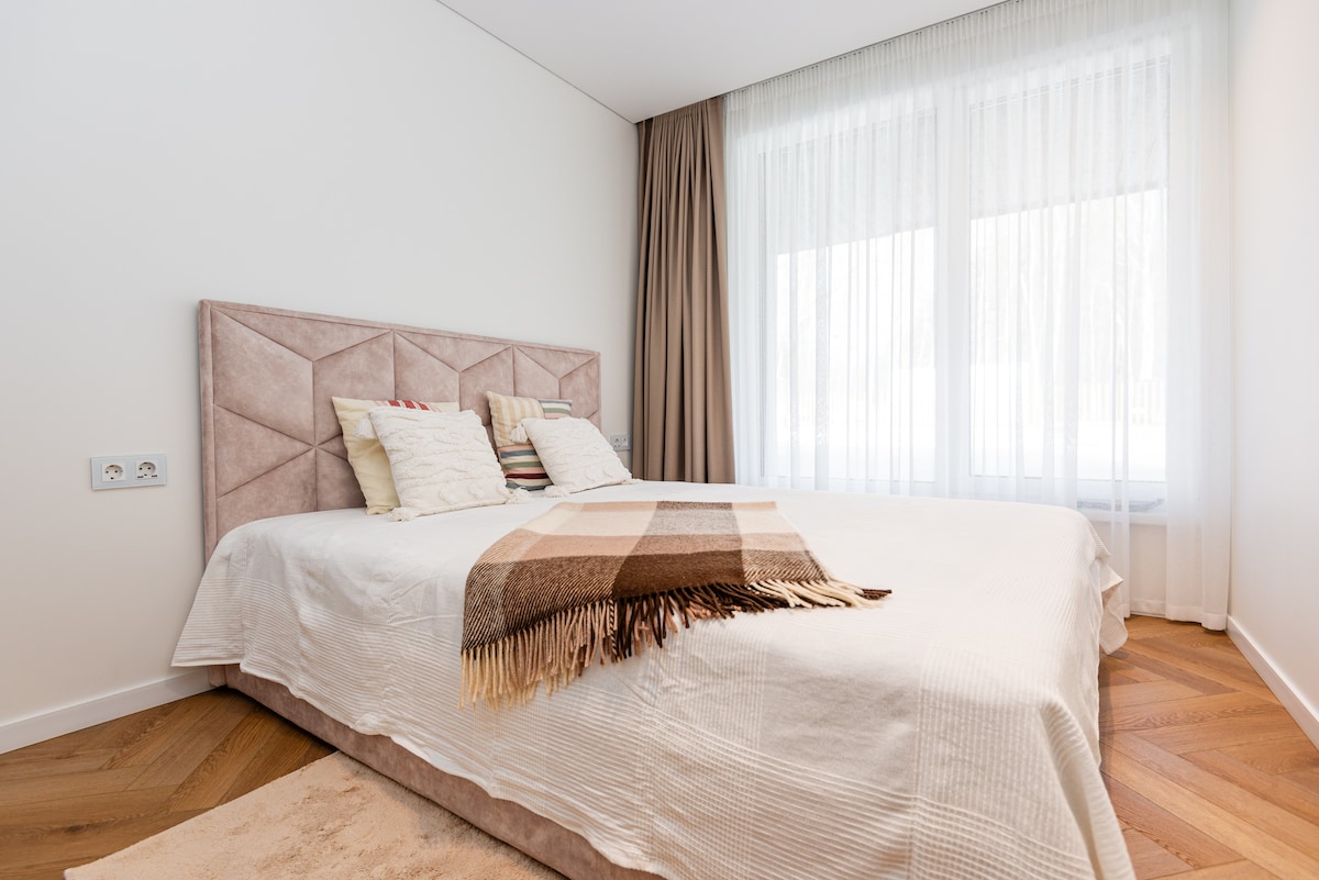 A spacious sleeping area features a soft, upholstered headboard and a neatly made bed with light bedding. Natural light filters through sheer curtains, illuminating the room. A textured throw blanket is draped across the foot of the bed, complementing the warm wood flooring.