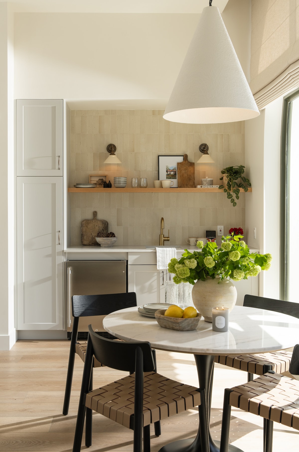 A modern kitchenette is showcased, featuring sleek cabinetry and stainless steel appliances. A circular dining table with black chairs is complemented by a vase of green flowers. Natural light from large windows enhances the clean and stylish design of the space.