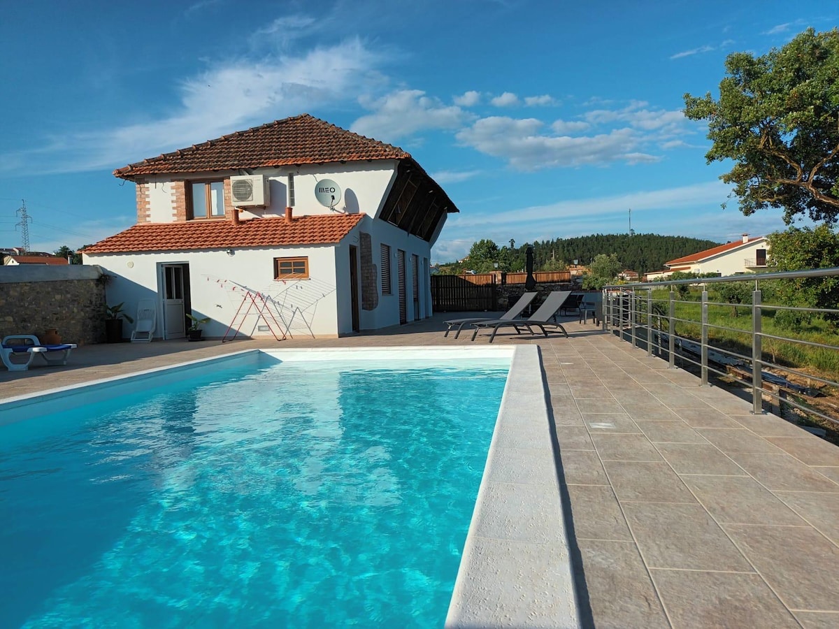 A seasonal outdoor pool reflects the sky, framed by a tiled deck. Sun loungers are positioned nearby, and a house with multiple air conditioning units stands in the background, showcasing a sloped roof and adjacent greenery.