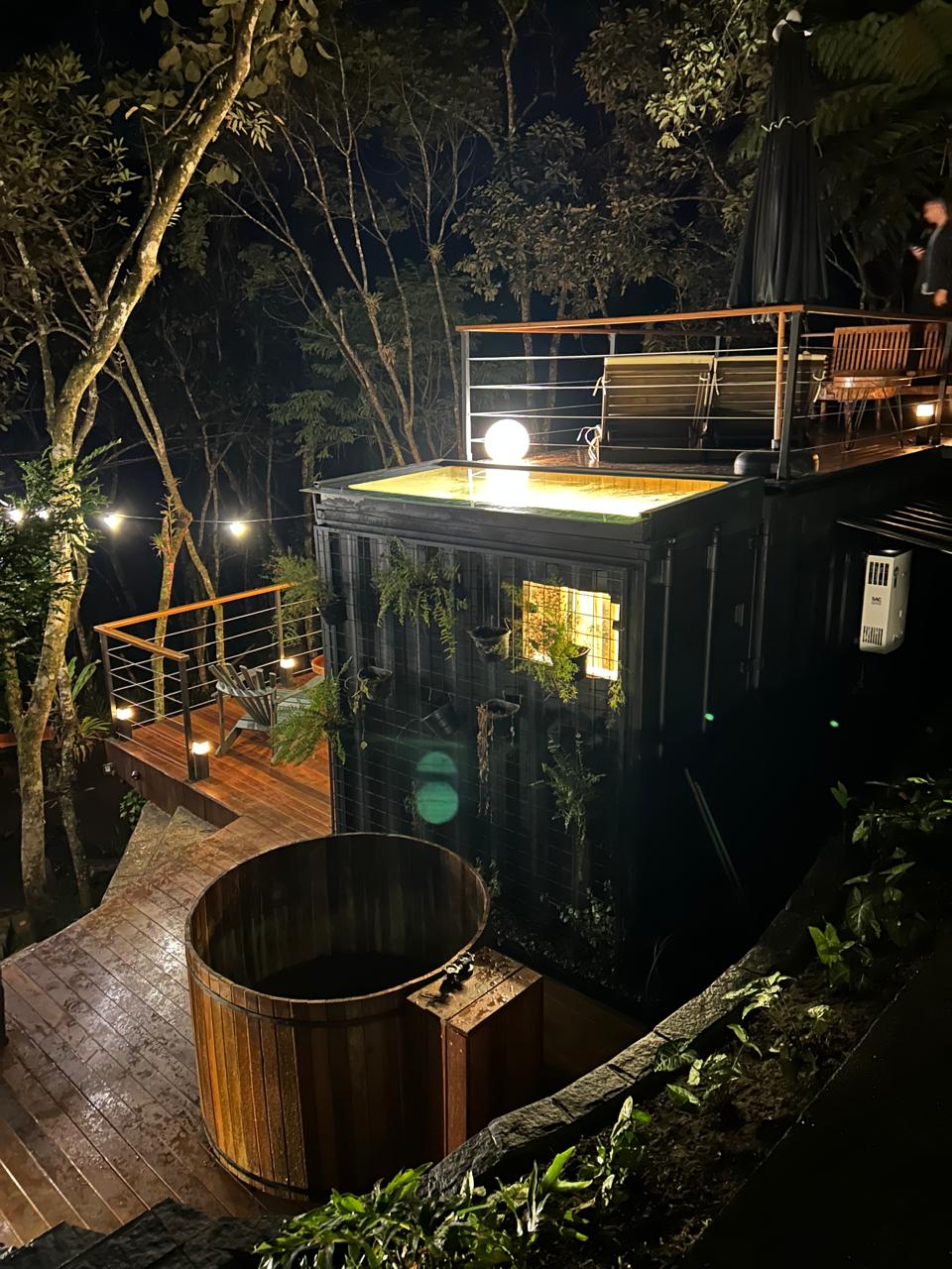 The image captures the exterior of a cabin at night, surrounded by trees. Soft lighting illuminates the upper deck, which features seating and a jacuzzi. A wooden structure, possibly a hot tub, is positioned nearby, enhancing the serene atmosphere of the natural setting.