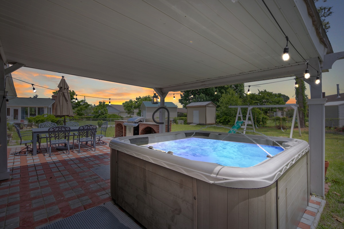 A spacious patio area features a large hot tub illuminated with soft lights, accommodating five to six people. Nearby, an outdoor dining table with an umbrella is positioned alongside a grill area, with a swing set visible in the background.