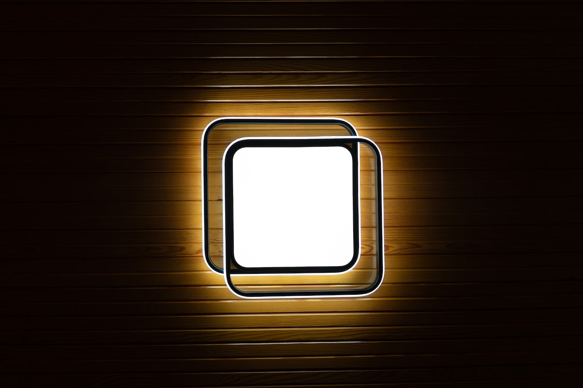 A modern light fixture is mounted on a wooden wall. The design features two overlapping square frames, with a warm glow emanating from the center, creating a soft illumination effect.