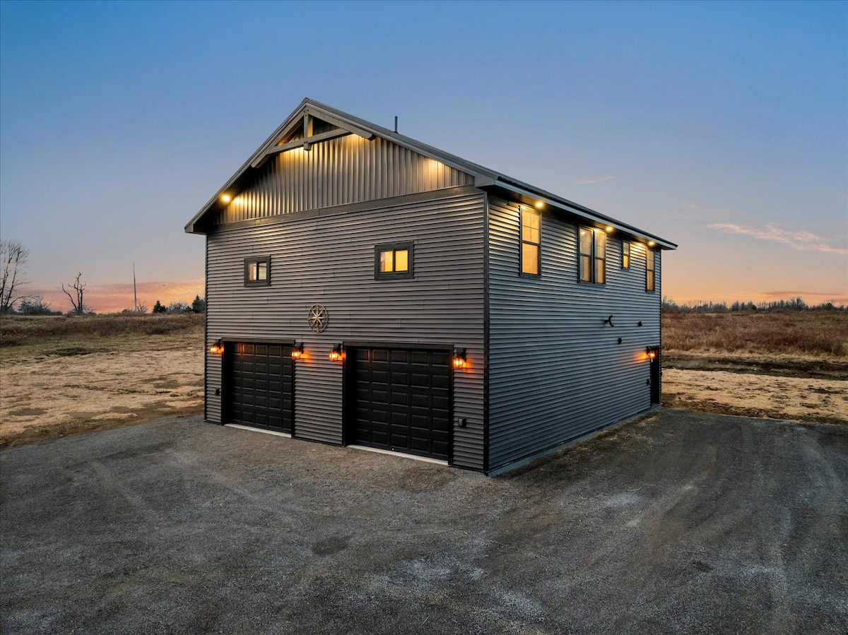 A modern two-story building is showcased, featuring a sleek metal exterior with illuminated garage doors. A spacious driveway leads to the front, while the surrounding land offers an open view. The structure is positioned against a twilight sky, enhancing its architectural appeal.