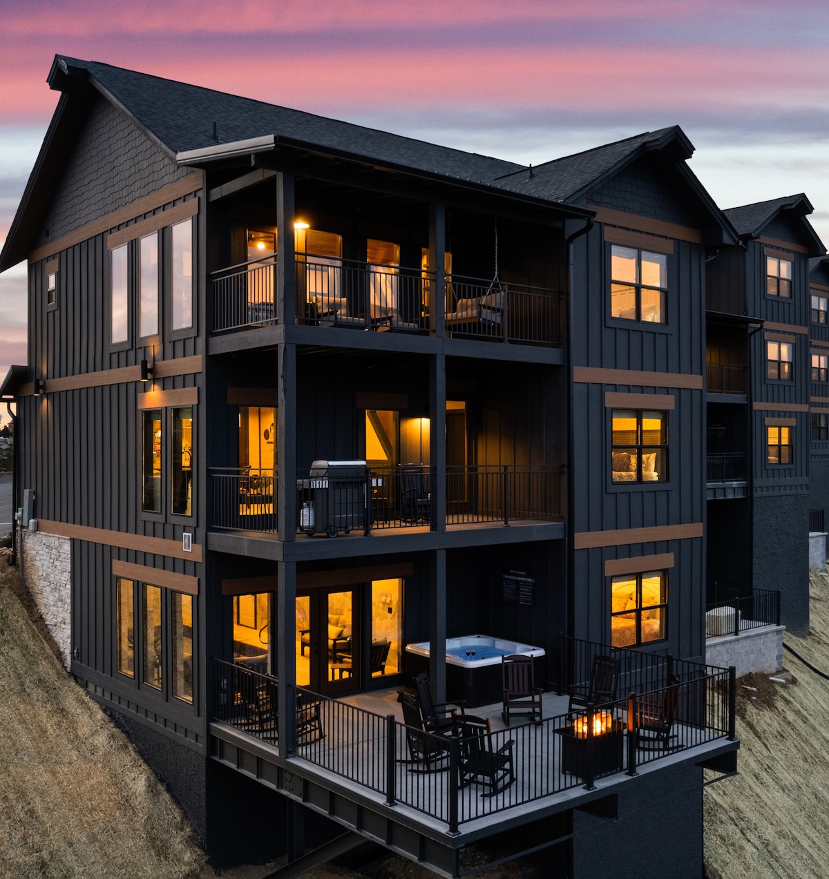 The multi-level cabin is set against a stunning backdrop, featuring dark exterior siding and large windows that glow warmly from within. Outdoor decks are equipped with seating and a hot tub, inviting guests to enjoy the serene surroundings and picturesque views.