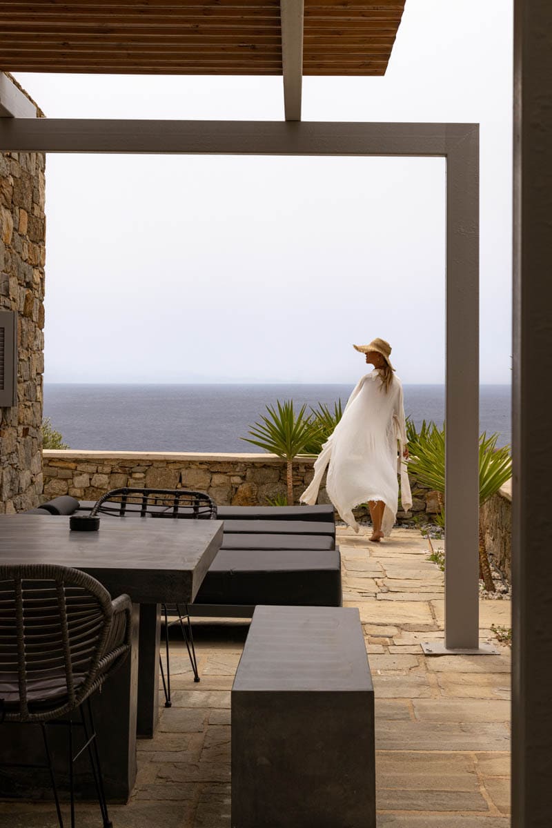An outdoor terrace is featured, with expansive views of the sea. Stone walls frame the area, complemented by dark dining furniture. Lush plants provide a touch of greenery, while the soft grey sky adds to the serene ambiance. A figure walks gracefully, dressed in light clothing.