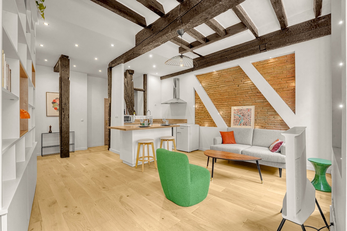 An open living area showcases wooden beams and a mix of textures. A green chair and gray sofa provide seating. The kitchen area features a wood countertop with seating for two. Soft lighting highlights a decorative wall art piece and vibrant accent pillows.