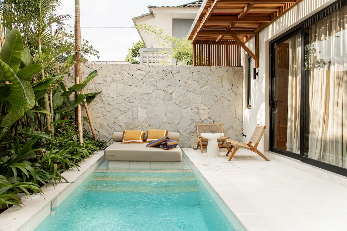 A serene private pool area features a modern design, surrounded by lush greenery. A spacious double sun bed sits on one side, complemented by a small table and a wooden chair. Large glass doors provide access to the villa's interior.