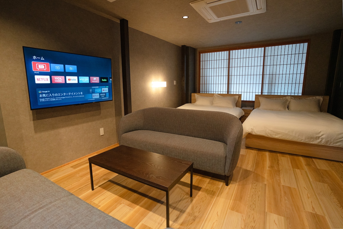 The guest room features a light wooden floor and a cozy seating area with a gray sofa. Two queen beds are positioned against the wall, separated by a narrow table. A wall-mounted smart TV is visible, providing access to streaming services, alongside traditional Japanese shoji windows allowing soft natural light.
