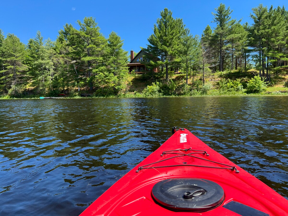 A vibrant red kayak floats on calm water, showcasing a view of a cottage nestled among tall pine trees. The clear blue sky reflects above, enhancing the tranquil atmosphere of Kennebec Lake.