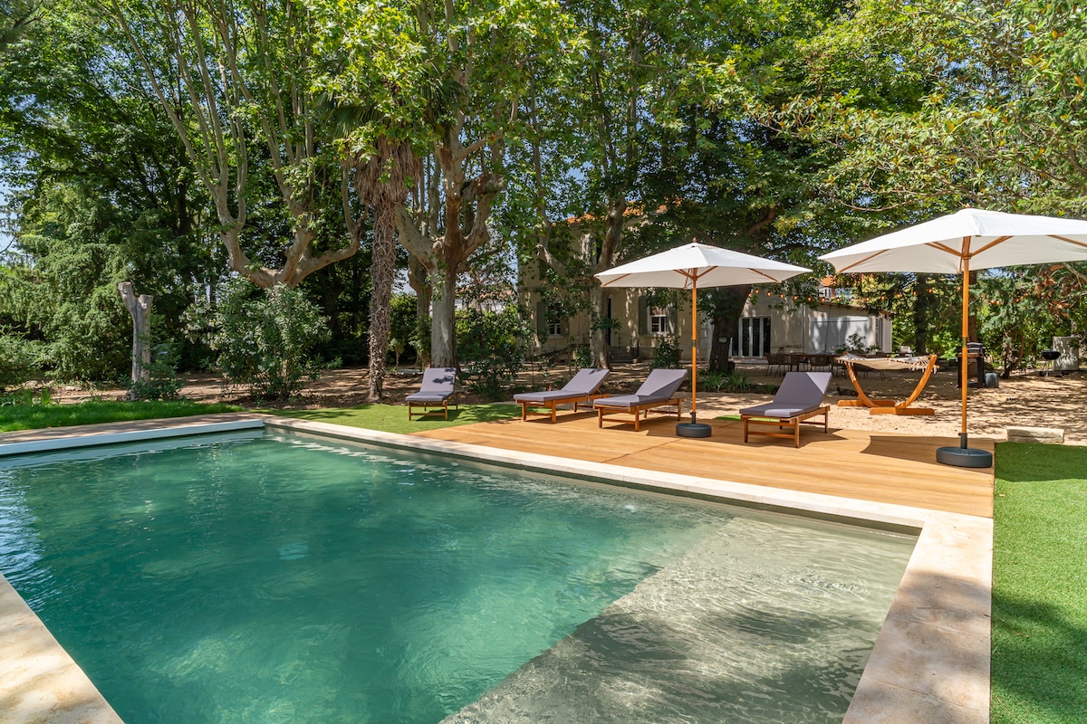 A serene pool area features a rectangular pool with clear water, surrounded by wooden decking. Sun loungers with cushions are positioned under large umbrellas, offering shaded relaxation. Lush greenery and trees provide a natural backdrop, enhancing the tranquil outdoor setting.