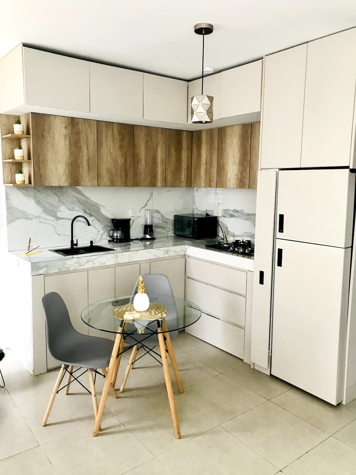 A modern kitchen design is displayed, featuring sleek cabinetry and a marble countertop. A small dining table with transparent chairs is positioned in the center. Kitchen appliances, including a refrigerator and coffee maker, are neatly arranged, contributing to the functional workspace.