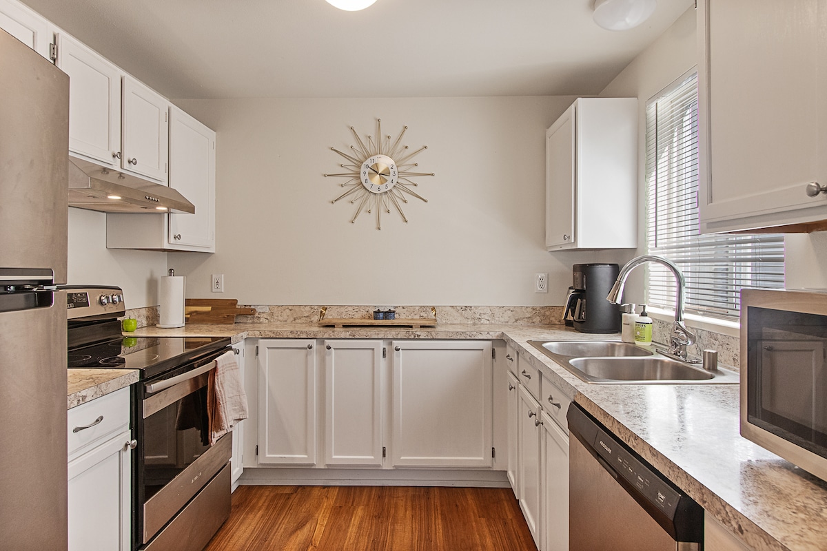 The kitchen features white cabinetry and a sunburst clock, complemented by stainless steel appliances. A spacious countertop allows for meal preparation, while natural light filters through the window. The room is organized with a coffee maker and microwave, enhancing functionality.