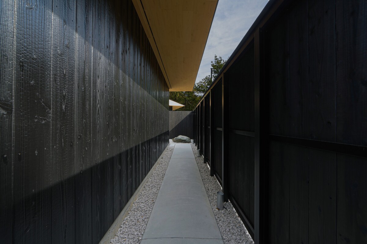 A narrow pathway is lined with dark wooden fences, leading through a landscaped area with pebbles on each side. The smooth concrete path extends towards a distant view of greenery beneath a softly illuminated sky.