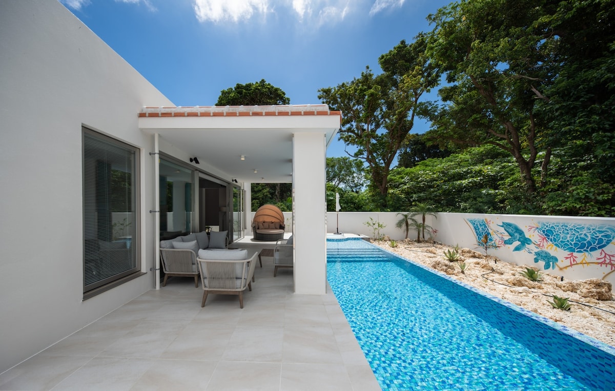 An outdoor area features a sparkling blue pool surrounded by tropical greenery. Comfortable seating arrangements are positioned under a covered patio, providing a space for relaxation. Sunlight filters through the trees, creating a serene ambience in this private setting.