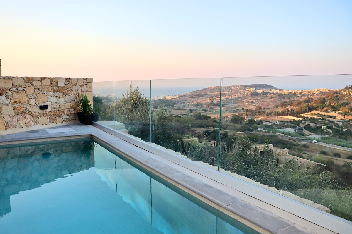 A clear view of a private pool surrounded by a glass railing, with serene valley landscapes stretching into the distance. The horizon displays soft pastel colors from the sunset, enhancing the tranquility of this outdoor space.