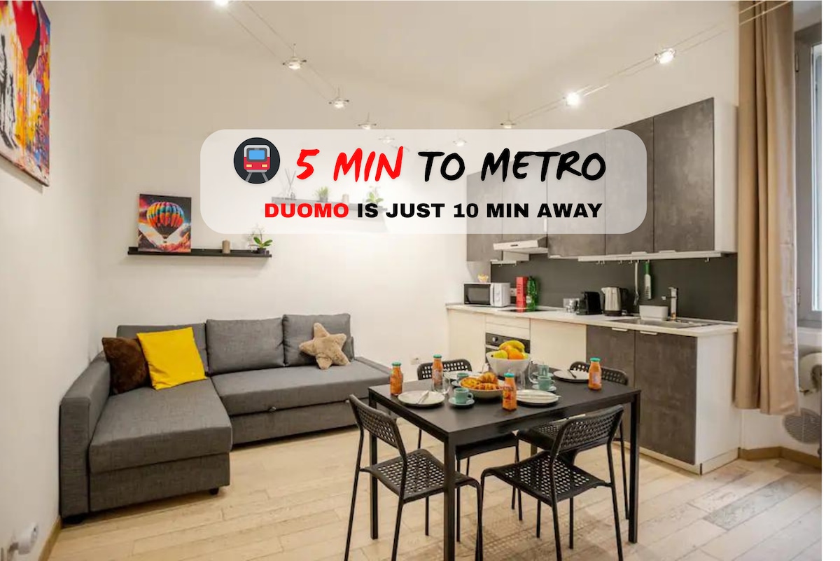 A modern living space features a comfortable gray sofa alongside a dining table set for four. Light wood flooring complements the neutral-colored walls, while overhead lighting adds brightness. A kitchenette is visible in the background, equipped with essential appliances for meal preparation.