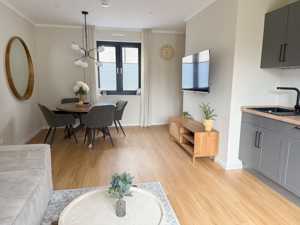 A modern living area features light wood flooring and a circular dining table surrounded by four upholstered chairs. A wall-mounted Smart-TV is positioned above a wooden console. Natural light enters through a large window, enhancing the neutral color palette.